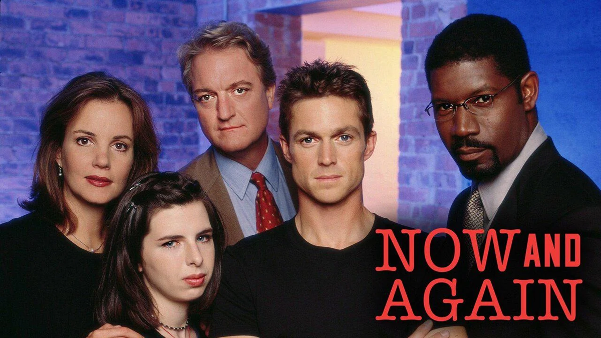 Five people pose together against a brick wall and blue background. Four have serious expressions, one smiles slightly. Red text in the lower right corner reads, “NOW AND AGAIN.”