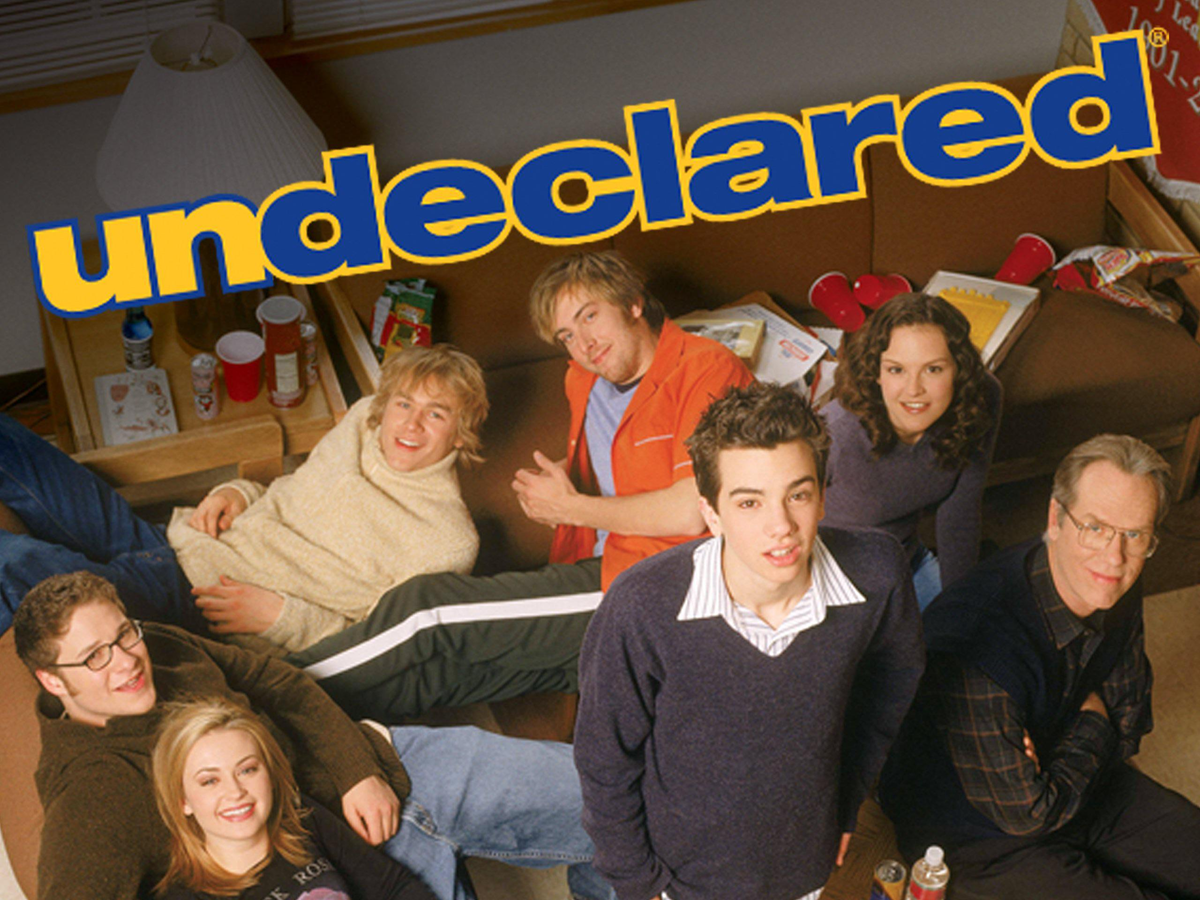 Seven young adults sit and stand around a couch in a casual room. The yellow and blue "undeclared" logo appears at the top. Cups and snacks are visible on a table near them.