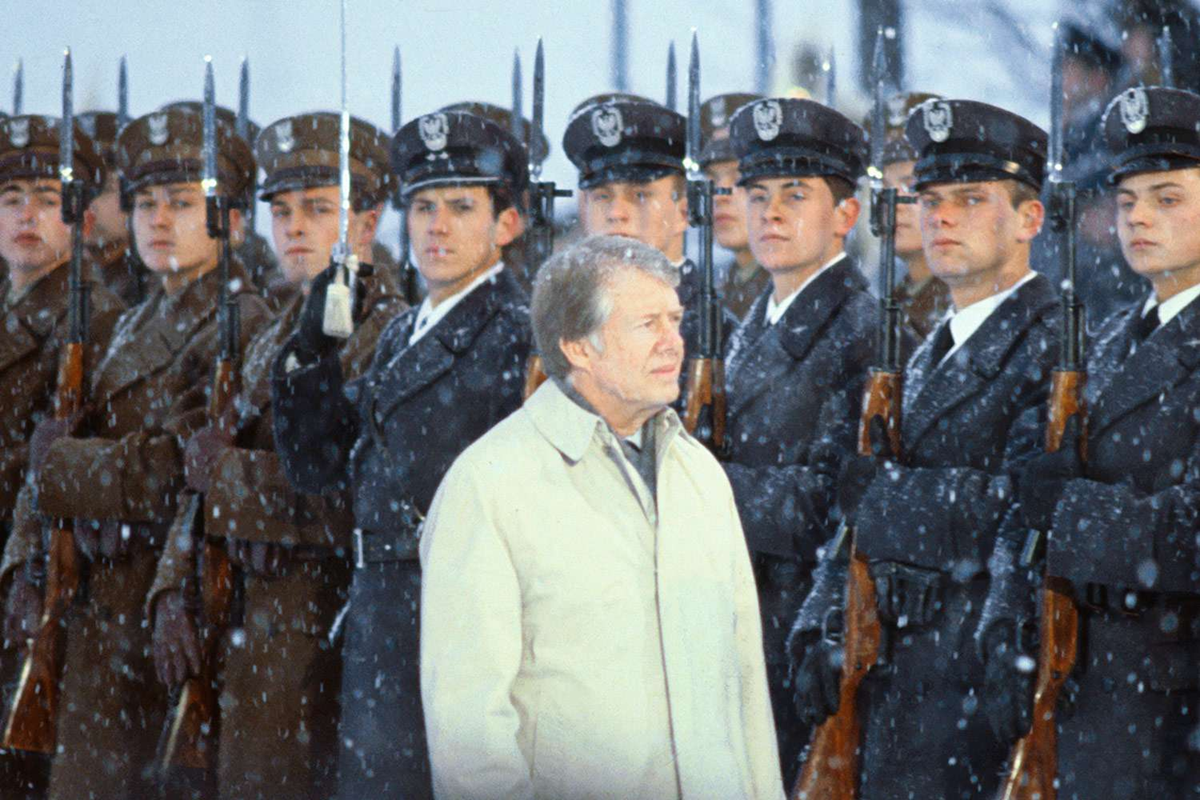 A man in a light coat stands outside in falling snow, surrounded by uniformed soldiers holding rifles upright, forming rows on both sides of him. The soldiers wear dark caps and serious expressions.