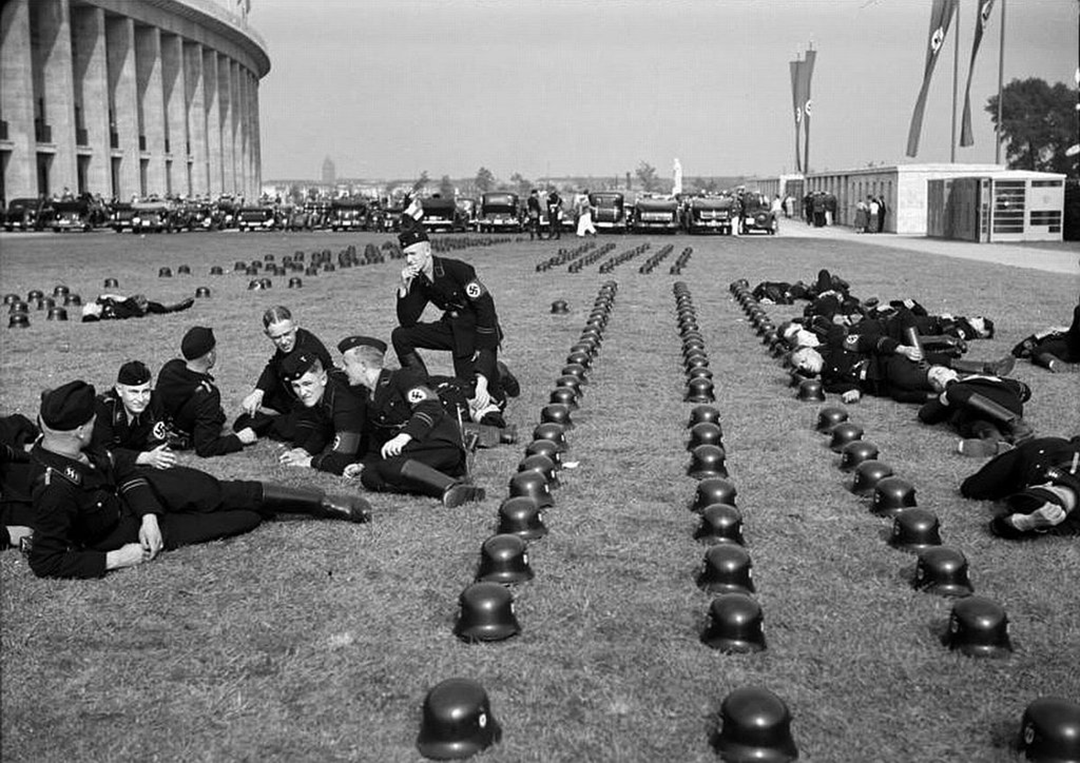 Groups of uniformed soldiers rest on grass beside rows of helmets near a large stadium; Nazi flags and vehicles are visible in the background.