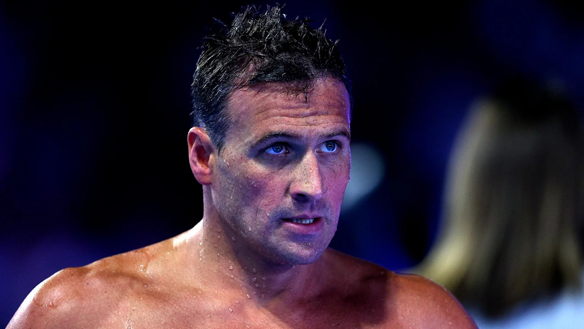 A male swimmer with wet hair and droplets on his shoulders looks ahead intensely, with a blurred figure and deep blue background behind him.