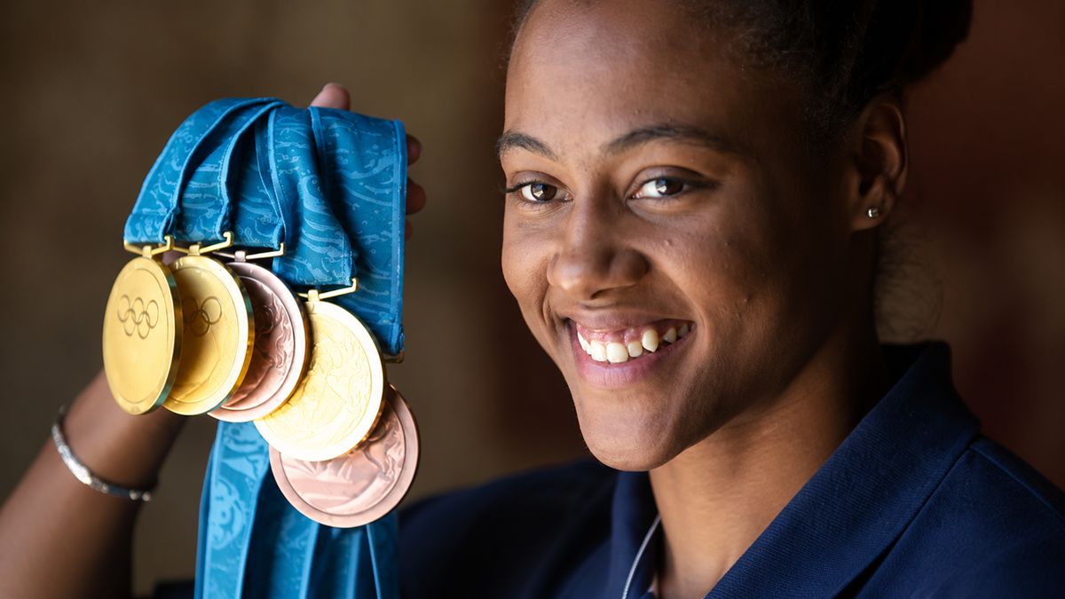 A smiling woman in a blue shirt holds up four Olympic medals on teal ribbons, showcasing them proudly.