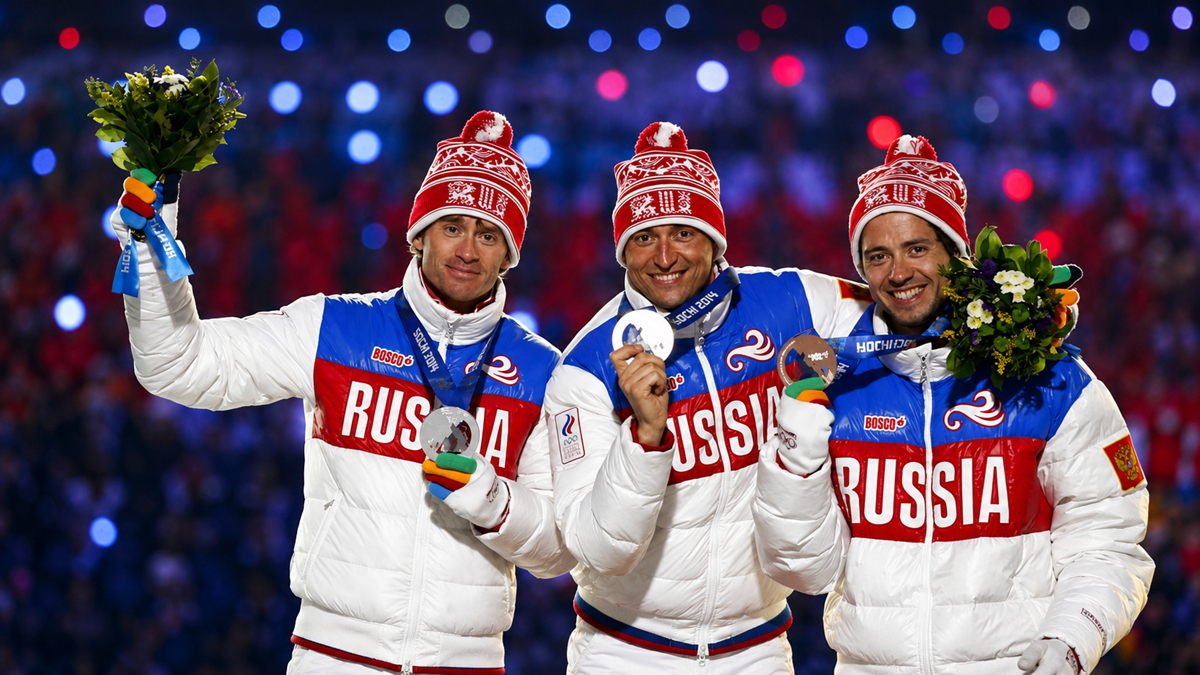 Three athletes in white "Russia" jackets and red hats hold medals and flower bouquets, smiling on a podium with a colorful, blurred crowd in the background.