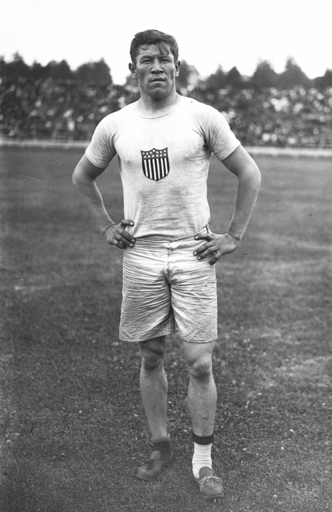 A male athlete stands on a field with his hands on his hips, wearing a white shirt with a USA shield emblem and light shorts; a crowd is visible in the background.