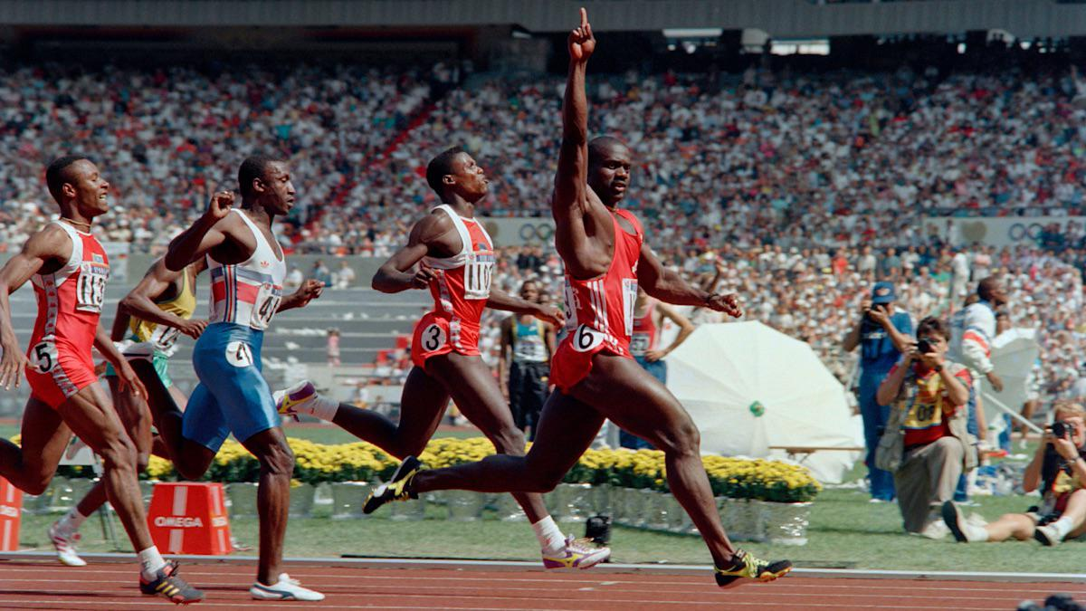 A sprinter in red raises his arm in victory as he crosses the finish line ahead of other runners on a track, with a large crowd and photographers in the background.