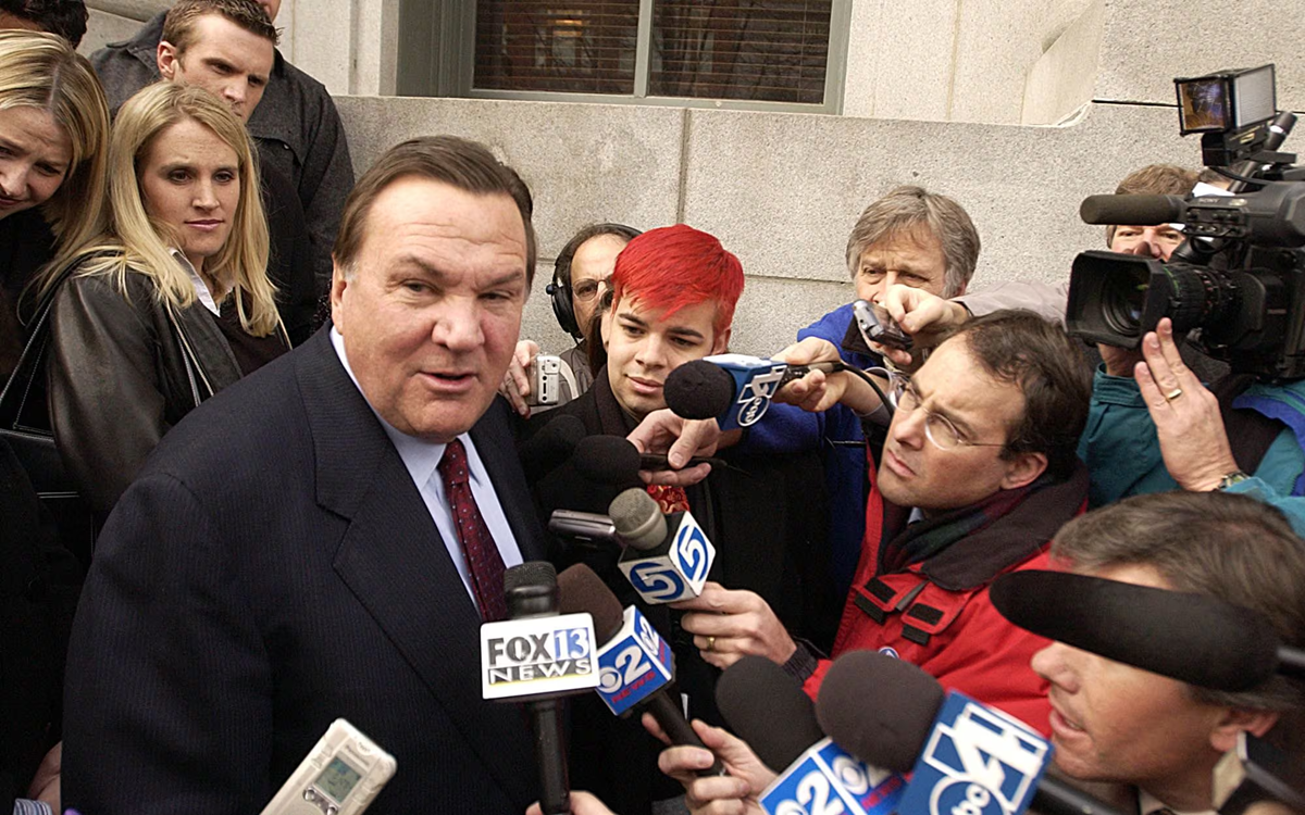 A man in a suit speaks to a crowd of reporters holding microphones and cameras outside a building, while people stand behind him listening and watching.