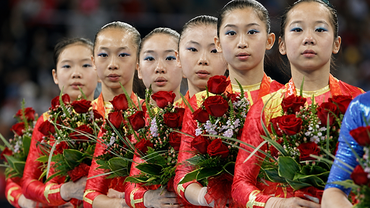 Six young female gymnasts in matching red uniforms hold bouquets of red roses, standing in a line and looking forward, during an award ceremony at a competition.
