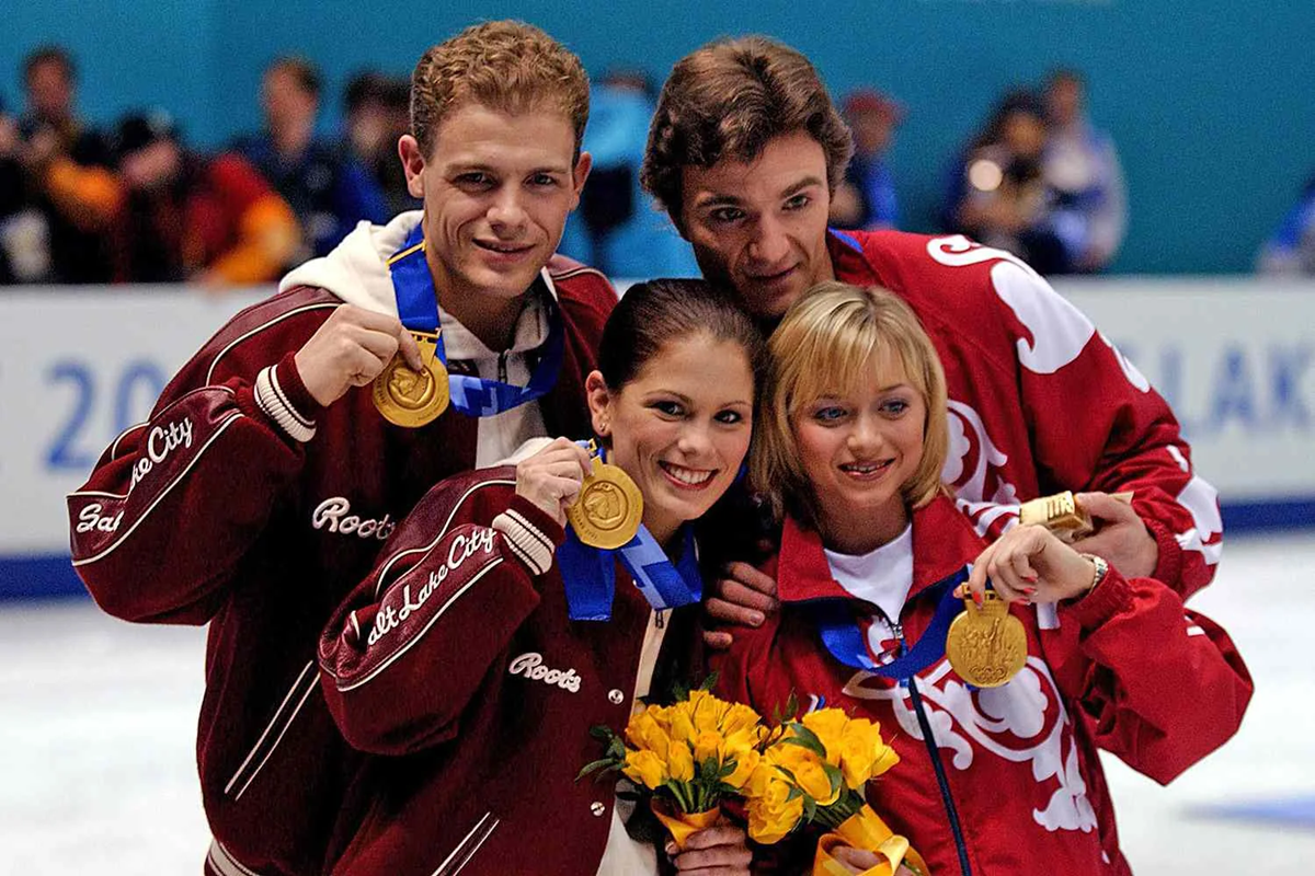 Four figure skaters pose on ice, each wearing a gold medal. Two hold bouquets of yellow flowers. All wear red and white jackets, smiling at the camera with a blue arena background and blurred crowd behind them.