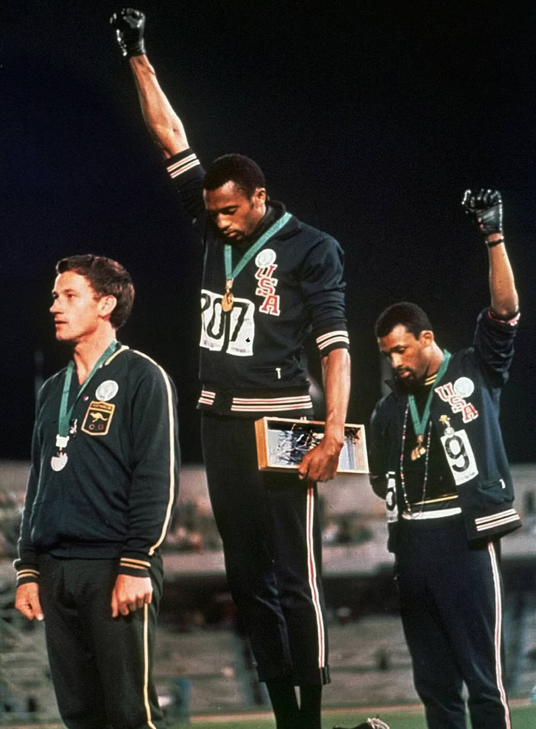 Three male athletes stand on an Olympic podium. Two, wearing USA jackets, raise black-gloved fists in protest. The third, from Australia, stands with arms at his sides.