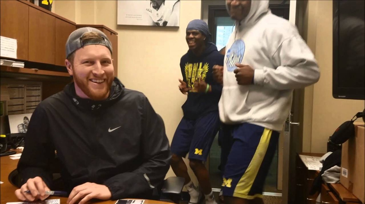 A man sits at a desk smiling, while two people in athletic gear dance joyfully behind him in an office setting.