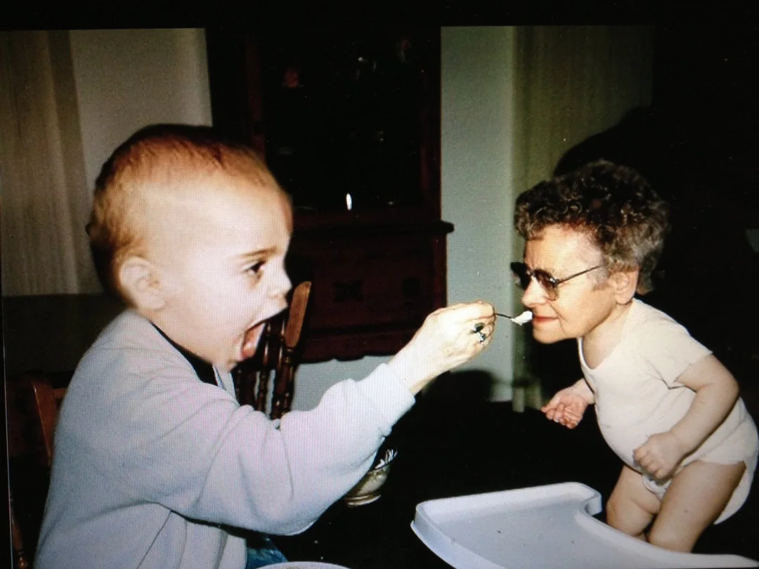 A child with an exaggeratedly open mouth pretends to feed a small person with an adult face, glasses, and a baby's body, sitting in a high chair. The scene is humorous and surreal.