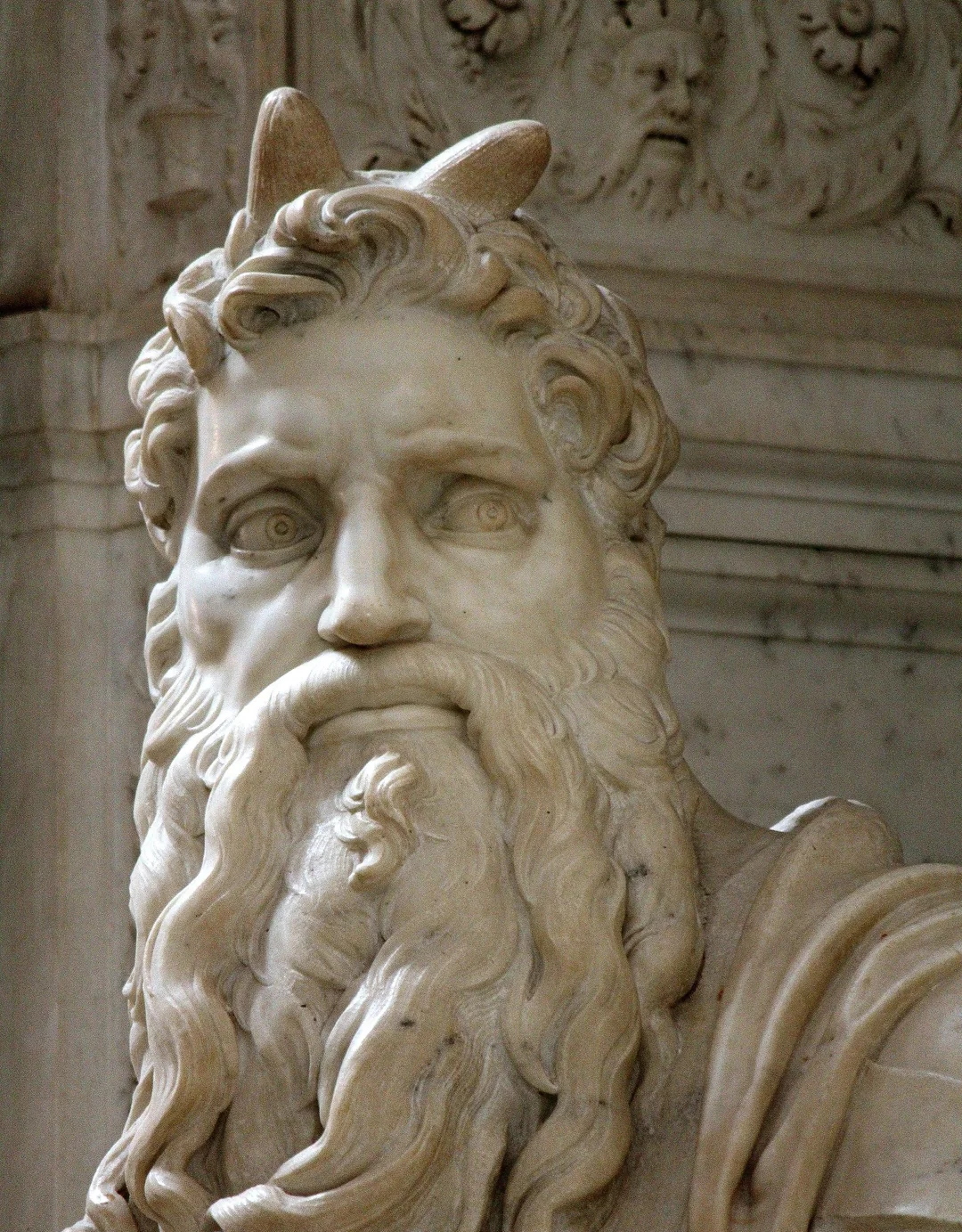 A close-up of a marble statue depicting a bearded man with flowing hair and two horns on his head, set against an ornate, carved background.