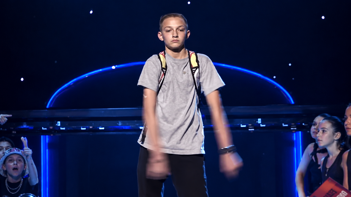 A young person in a gray shirt and backpack dances on stage with arms swinging, while a crowd watches. Blue lights and stars are visible in the background.