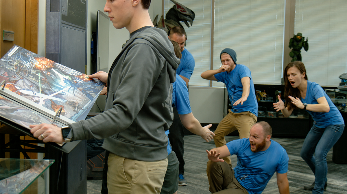 A person reads from a large book while five others react dramatically behind them, appearing surprised or alarmed. The setting is an office with large windows and pop culture decor. 2016