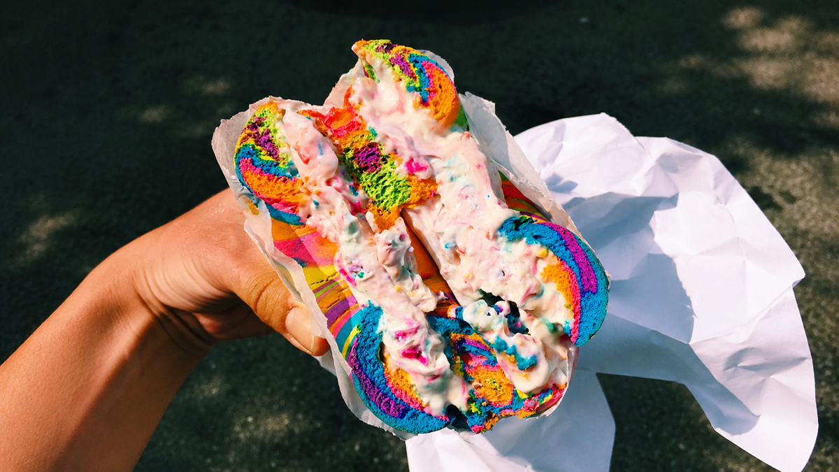 A hand holds a rainbow-colored bagel filled with sprinkled cream cheese, wrapped partially in white paper, with a dark, blurred background.