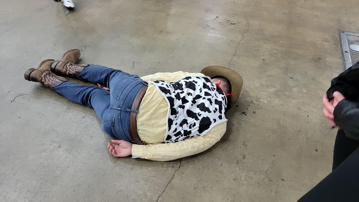 A person dressed as a cowboy, wearing a cow-print vest and boots, lies face down on a concrete floor with hands behind their back. Others stand nearby.