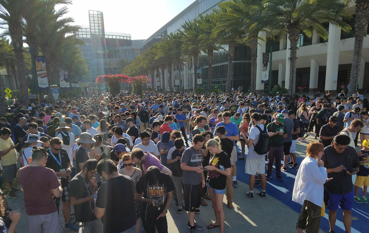 A large crowd of people stands outside a building lined with palm trees, many looking at phones. The scene is sunny, and the group appears to be waiting for an event or gathering outdoors.