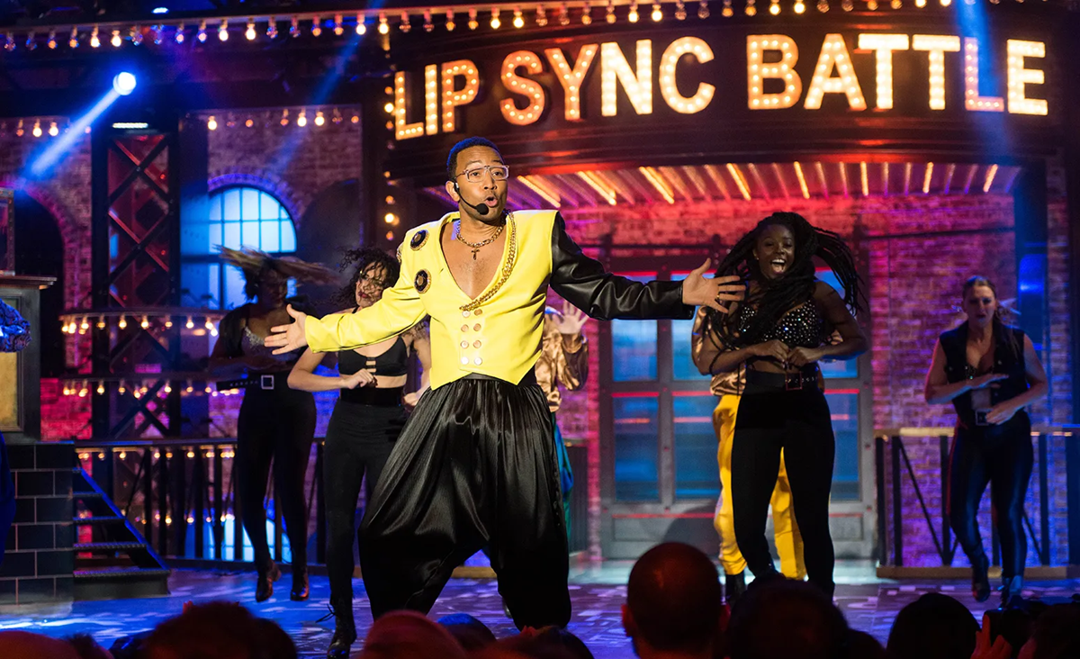 A performer in a yellow and black costume energetically dances on stage with backup dancers, under large illuminated "Lip Sync Battle" sign.