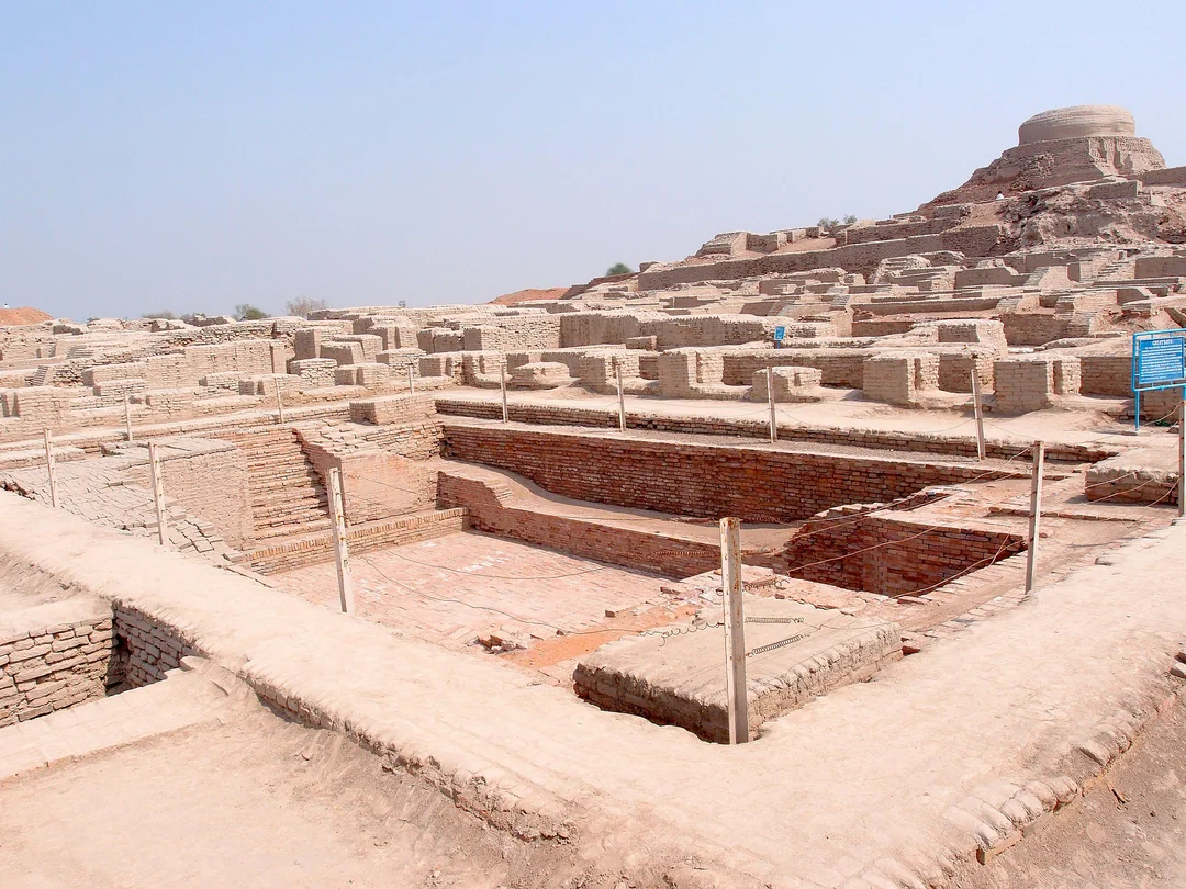 Ancient brick ruins of Mohenjo-daro under a clear sky, featuring rectangular foundations, walls, and a large structure in the background, with some ropes marking pathways around the site. Civilization.