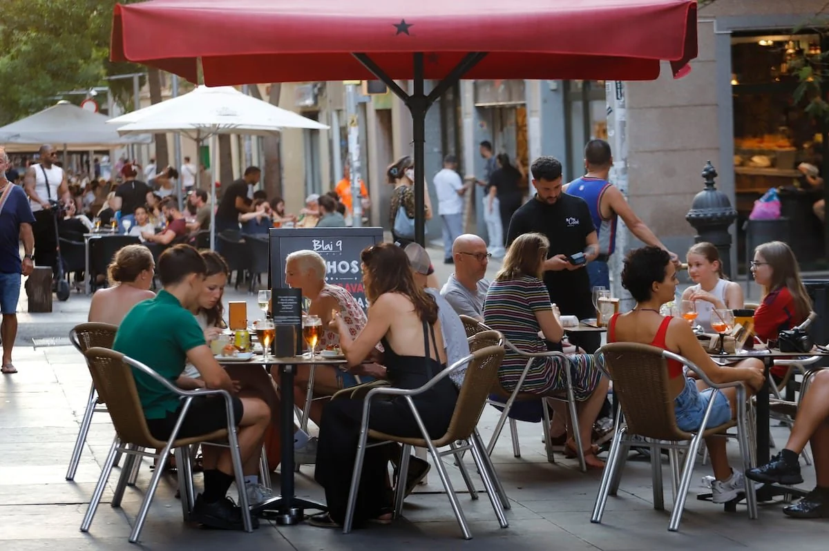 People sit at outdoor café tables under umbrellas on a busy street, enjoying food and drinks. A waiter serves a table while pedestrians walk by and other diners relax in the background. It is a lively, social atmosphere.