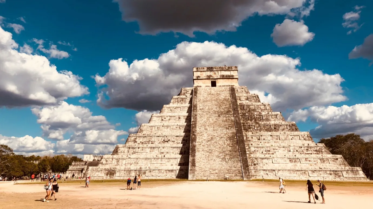 A large stone pyramid, known as El Castillo at Chichen Itza, stands under a partly cloudy sky with tourists walking around its base on a sunny day.
