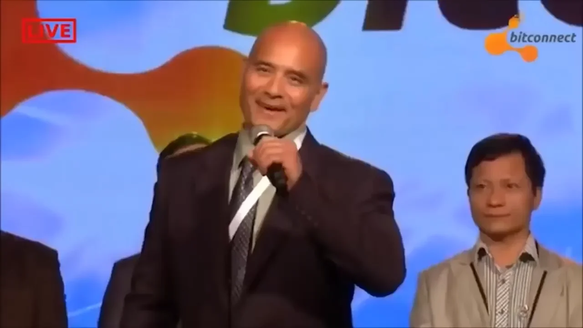 A man in a suit speaks into a microphone on stage with a colorful background and two other people partially visible behind him.