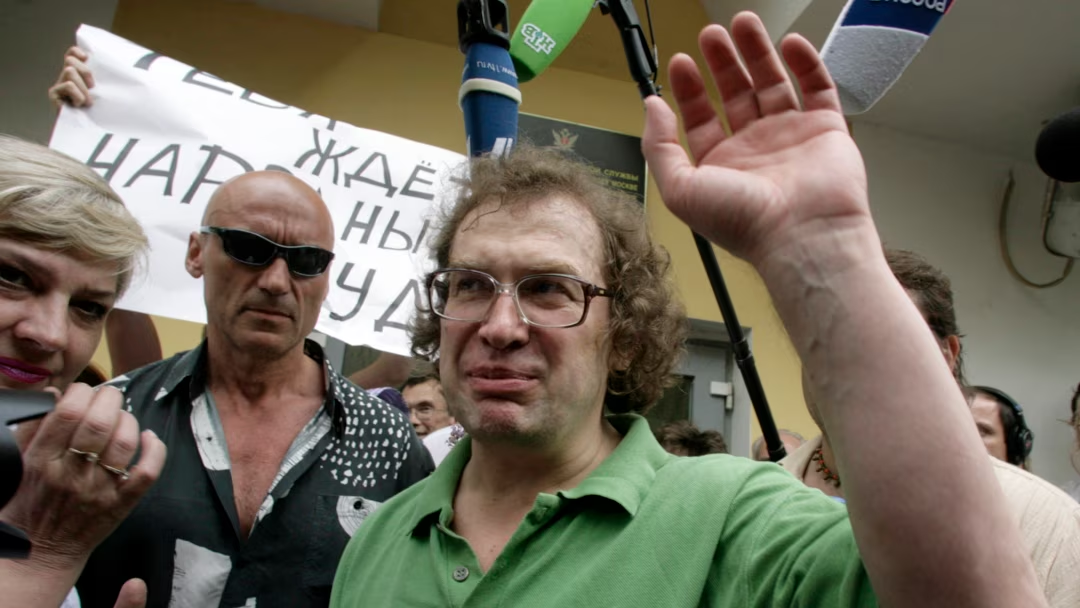 A man in a green shirt raises his hand while surrounded by people and microphones. A banner with Cyrillic text is visible in the background. The scene appears to be a public gathering or event.