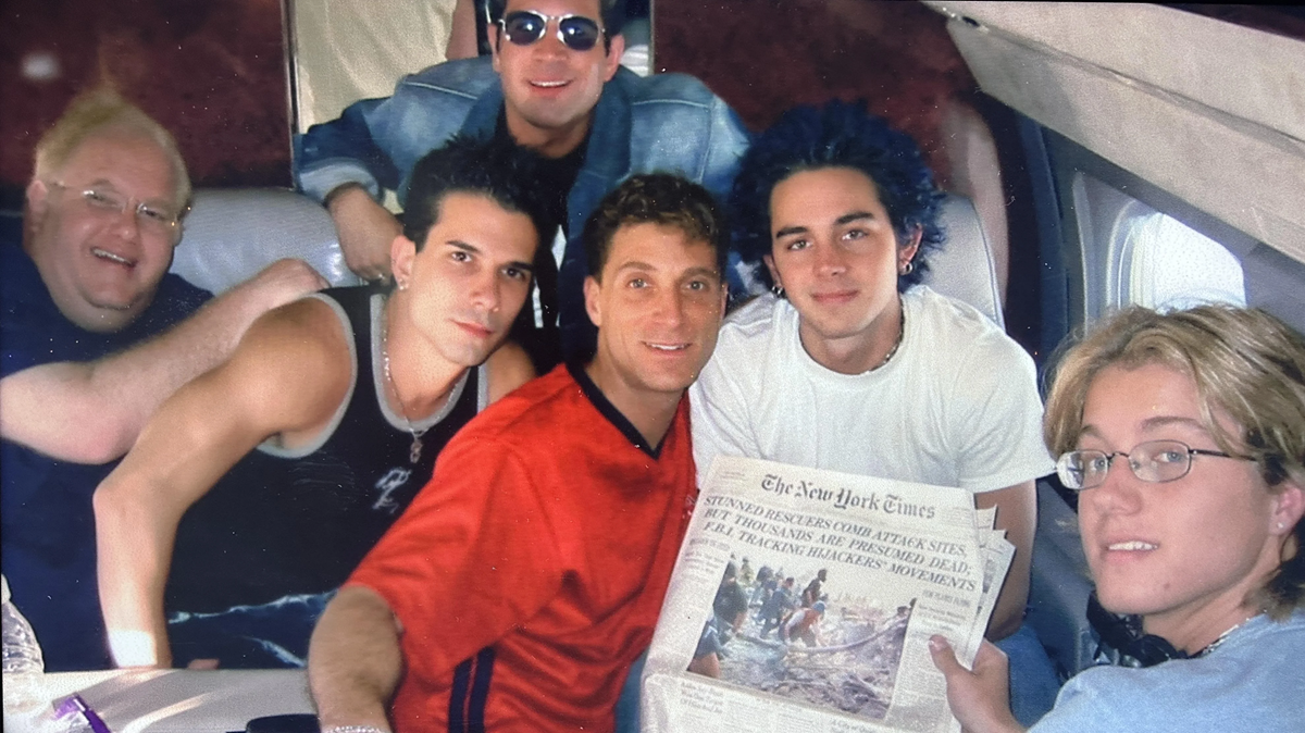 Six people sit together in a private jet, smiling at the camera. One person in front holds a copy of The New York Times newspaper. The group appears relaxed and casual, dressed in informal clothes.