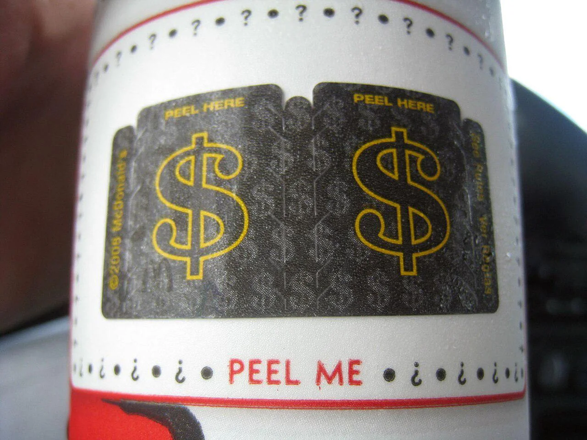 Close-up of a label with two peel-off black tabs featuring yellow dollar signs and the words "PEEL HERE." Below, red text says "PEEL ME.