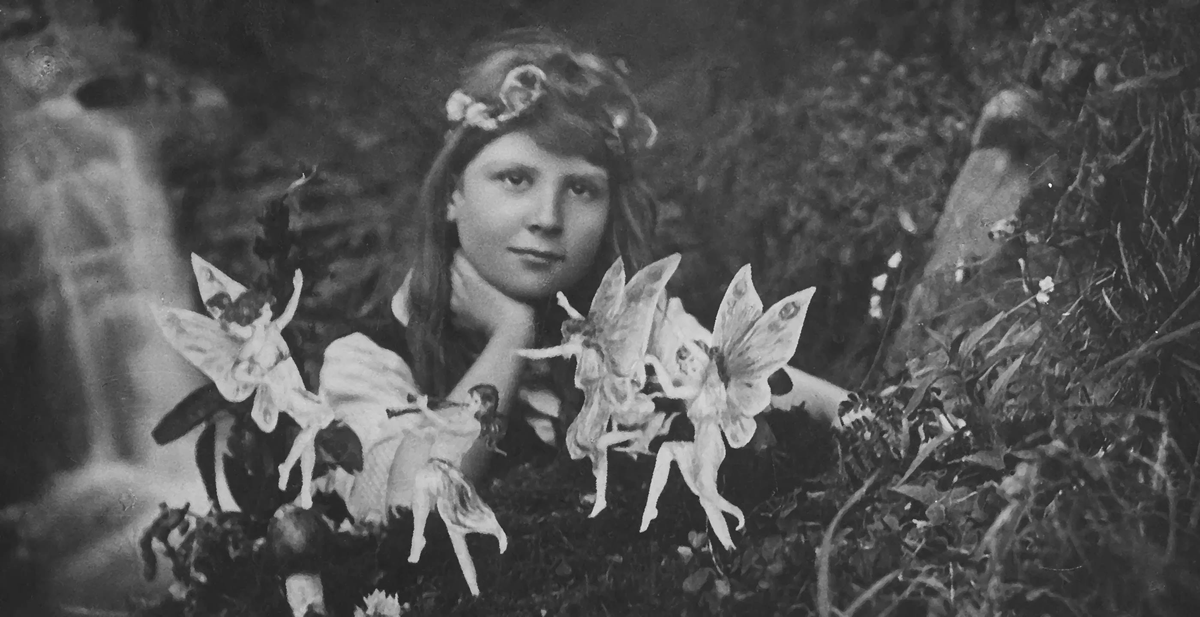A girl with a flower crown poses outdoors, surrounded by cut-out paper fairies among foliage, in a black-and-white, vintage-style photo.