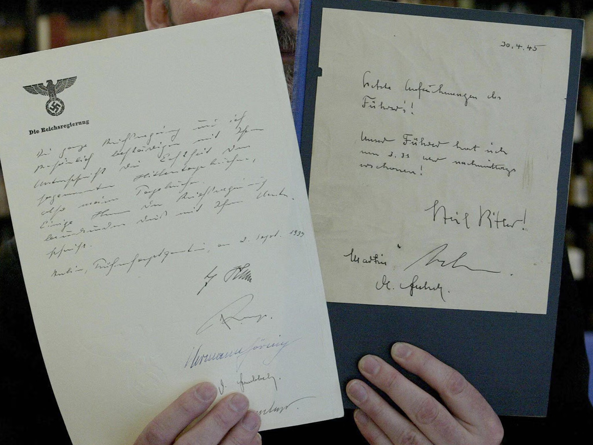 Two hands hold up two old handwritten German documents with signatures. The left paper features a Nazi eagle and swastika symbol at the top. The right paper has a date "20.12.45" in the corner. Both papers are yellowed with age.