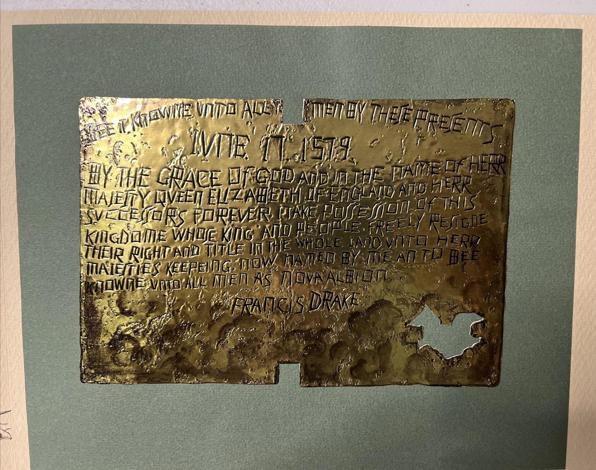 A weathered brass plaque with engraved, uneven text, dated June 17, 1579, referencing Queen Elizabeth and Francis Drake. The plaque is mounted on green paper and has some holes and surface damage.