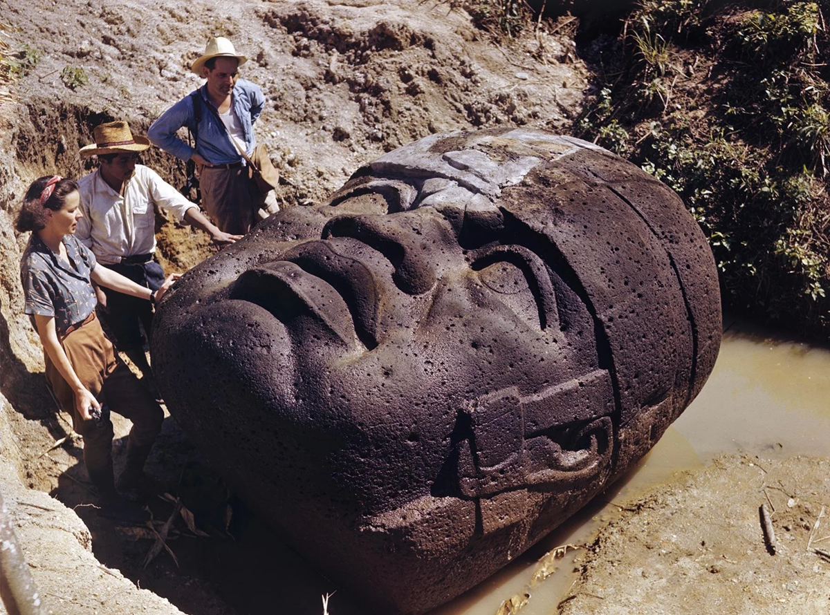 Three people stand around a large Olmec stone head partially buried in the ground, examining the ancient sculpture in an outdoor archaeological excavation site.