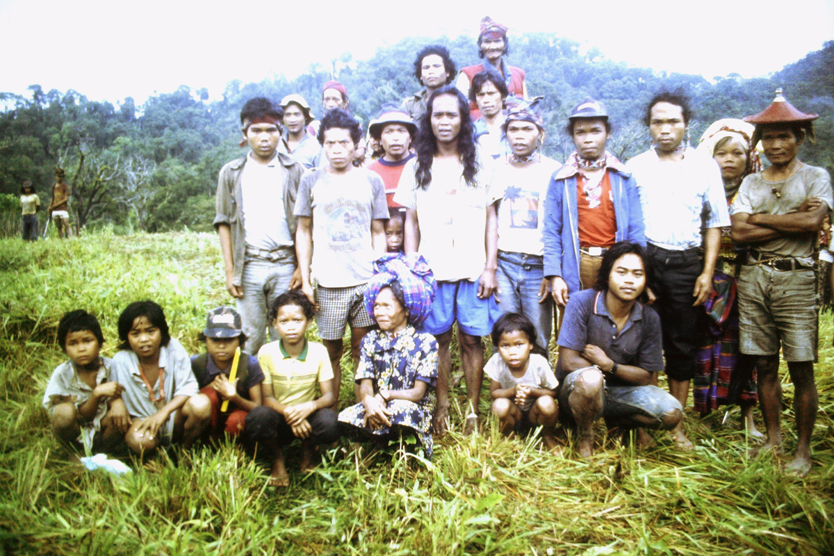 A group of people pose outdoors in a grassy field with trees and hills in the background. Some are standing, others are seated on the grass, and most are facing the camera.