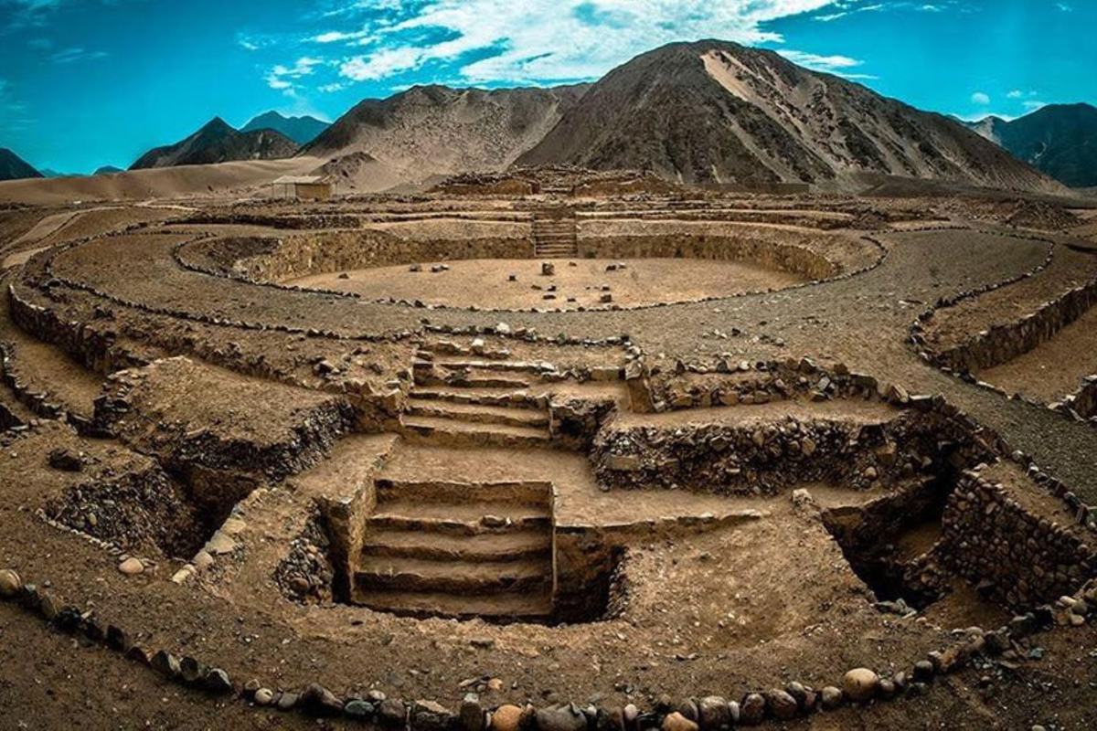 Ancient stone ruins with circular and rectangular formations and stairs, set in a dry, mountainous landscape under a blue sky with scattered clouds.