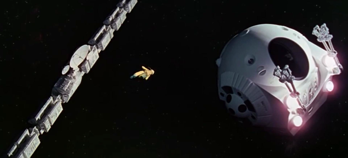 An astronaut floats in space between a long spacecraft and a round space pod, against a black starless background.