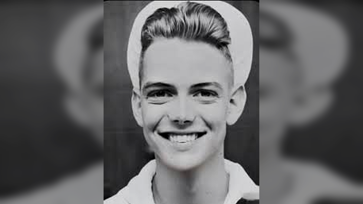 A young man in a sailor uniform smiles at the camera. He has short, neatly styled hair and is wearing a white sailor cap. The image is black and white.
