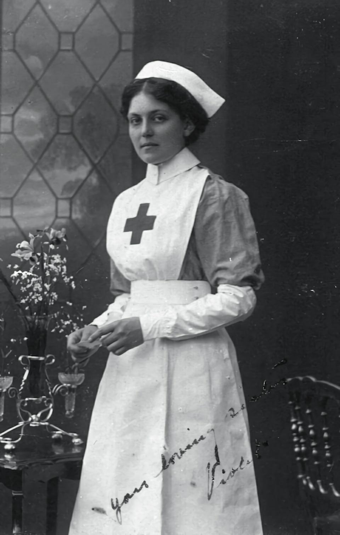 Black and white photo of a woman in a nurse’s uniform with a Red Cross emblem, standing beside a table with flowers, in front of a window and dark backdrop. Handwritten text appears at the bottom.