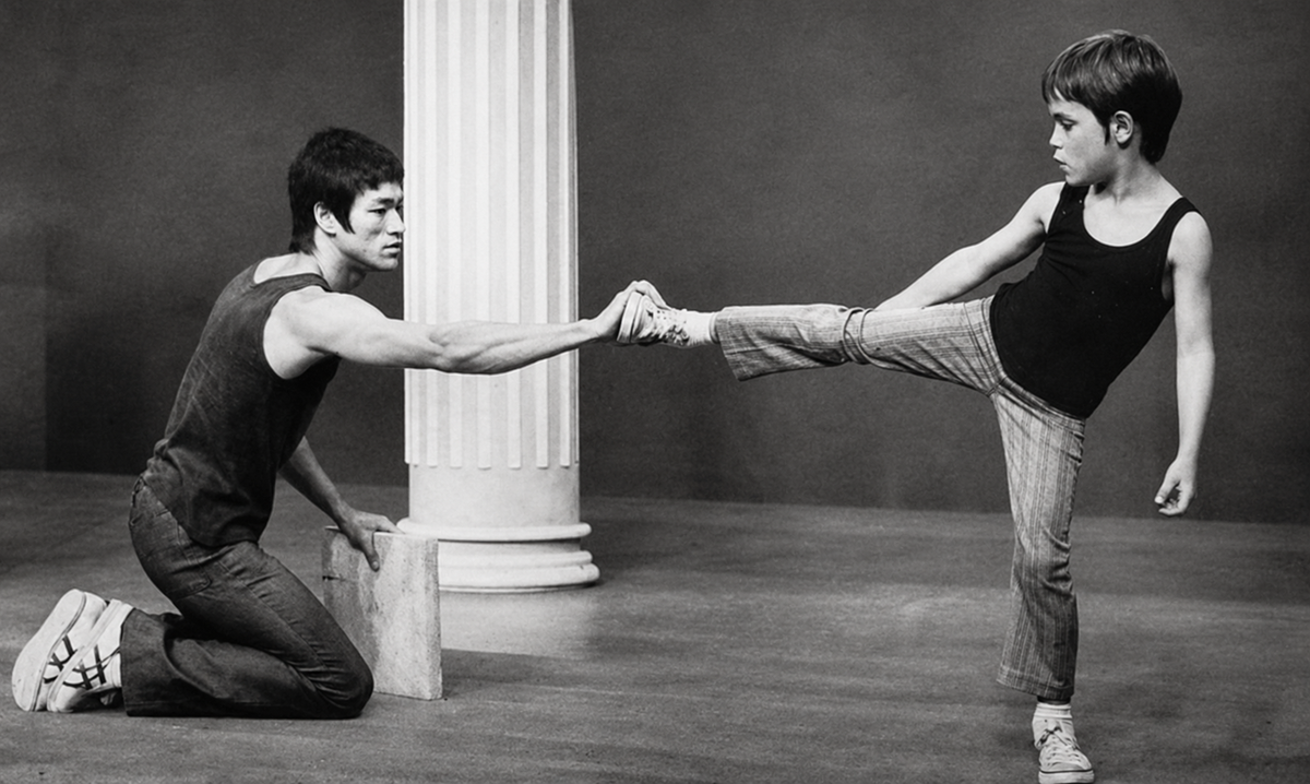 A man kneels and holds a young boy’s outstretched leg as the boy practices a high kick. Both wear casual clothes and stand in front of a white column.