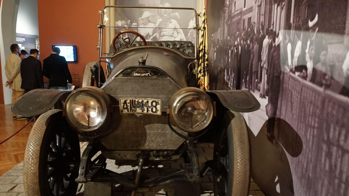 A vintage car with a worn license plate is displayed in a museum. People in early 20th-century attire are seen in a large black-and-white photo on the wall behind the car.