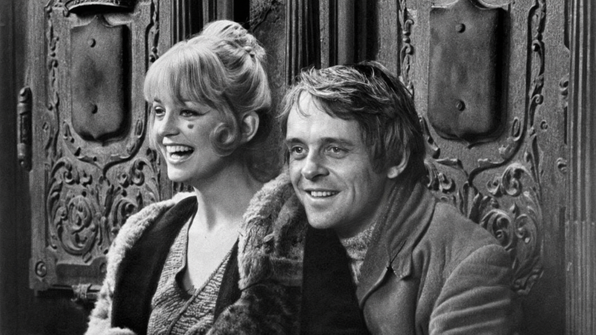 A smiling woman and man sit close together in front of an ornate door, wearing coats. The photo is black and white, and both appear happy and relaxed.