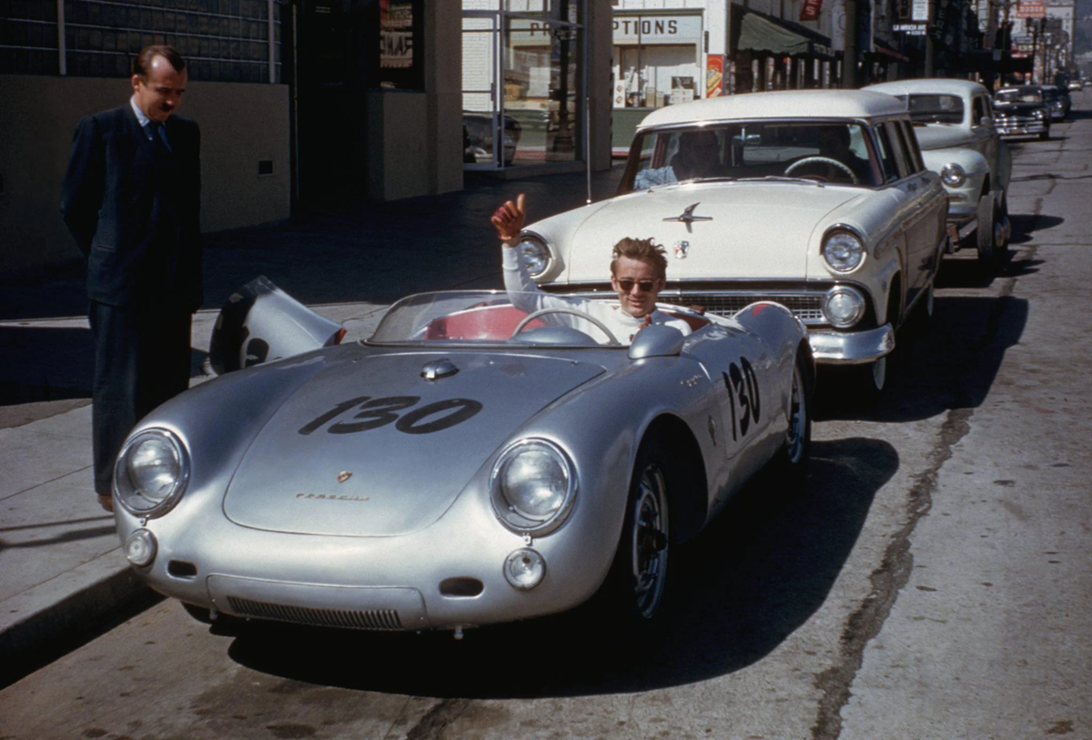 A man wearing sunglasses sits in a silver Porsche 550 Spyder with the number 130, parked on a city street. Another man stands on the sidewalk. A white classic car is parked behind them.