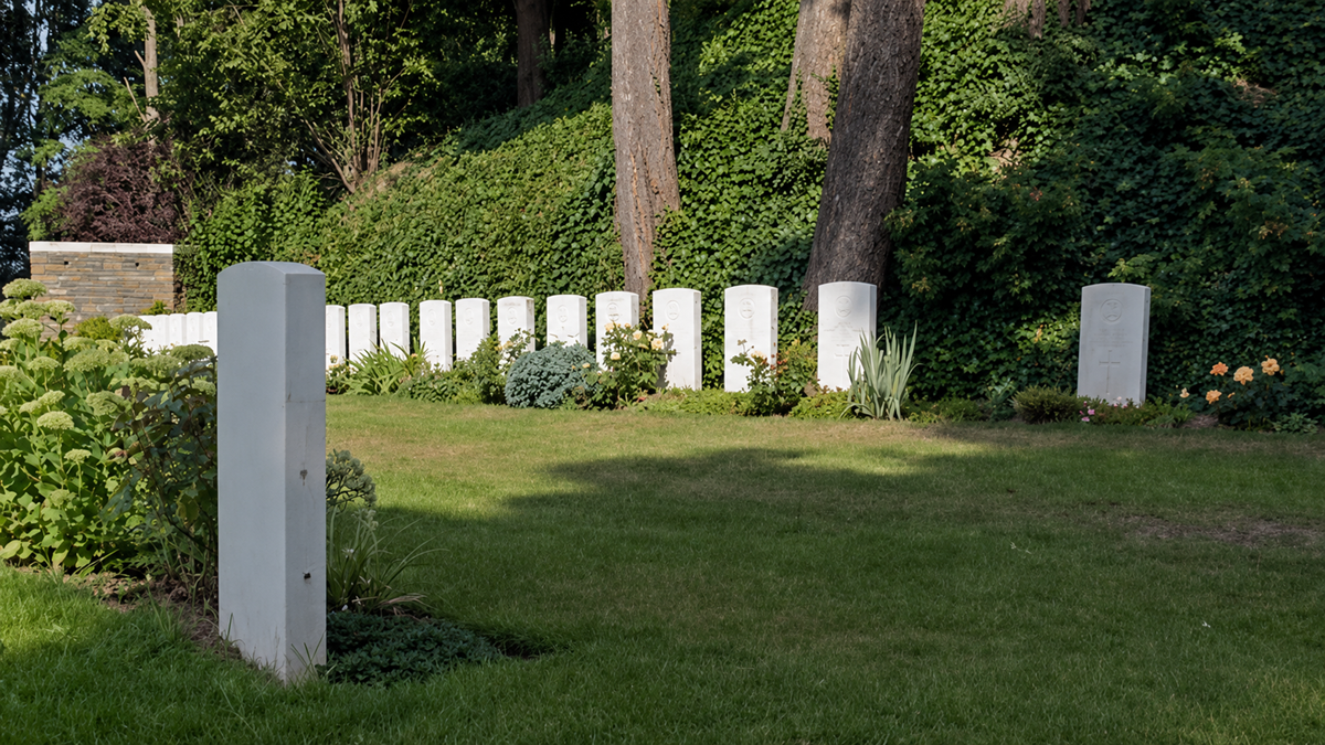 Rows of white gravestones stand on a green lawn surrounded by lush plants and tall trees, forming a serene and peaceful cemetery scene.