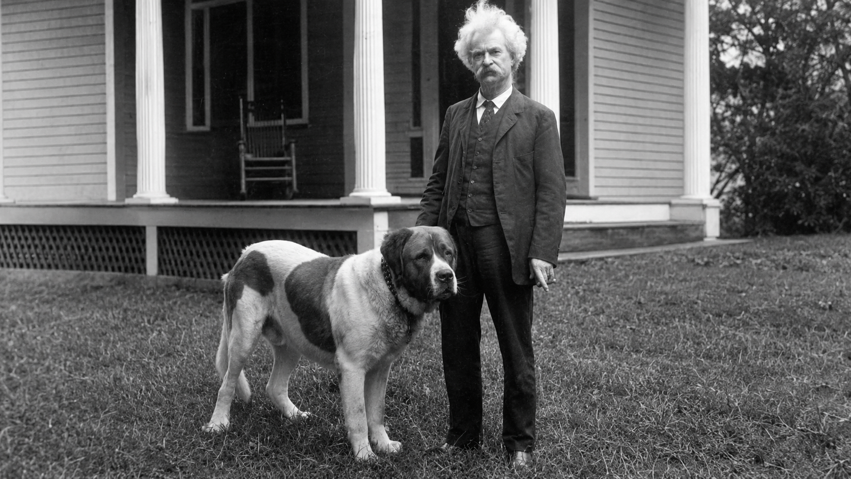 An older man in a suit stands beside a large St. Bernard dog on grass in front of a porch with columns and a wooden rocking chair. Coincidence.