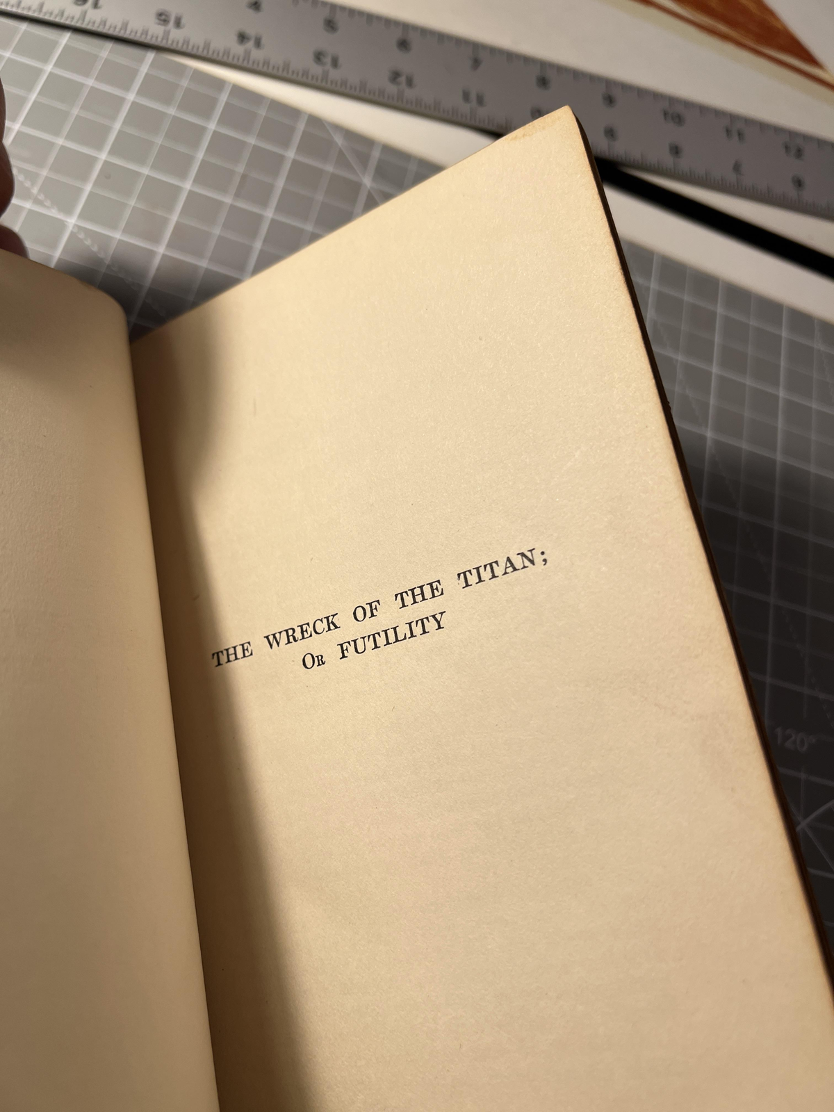 An open book shows the title page with the words "THE WRECK OF THE TITAN; Or FUTILITY" in typewriter font, with a ruler and a cutting mat visible in the background.