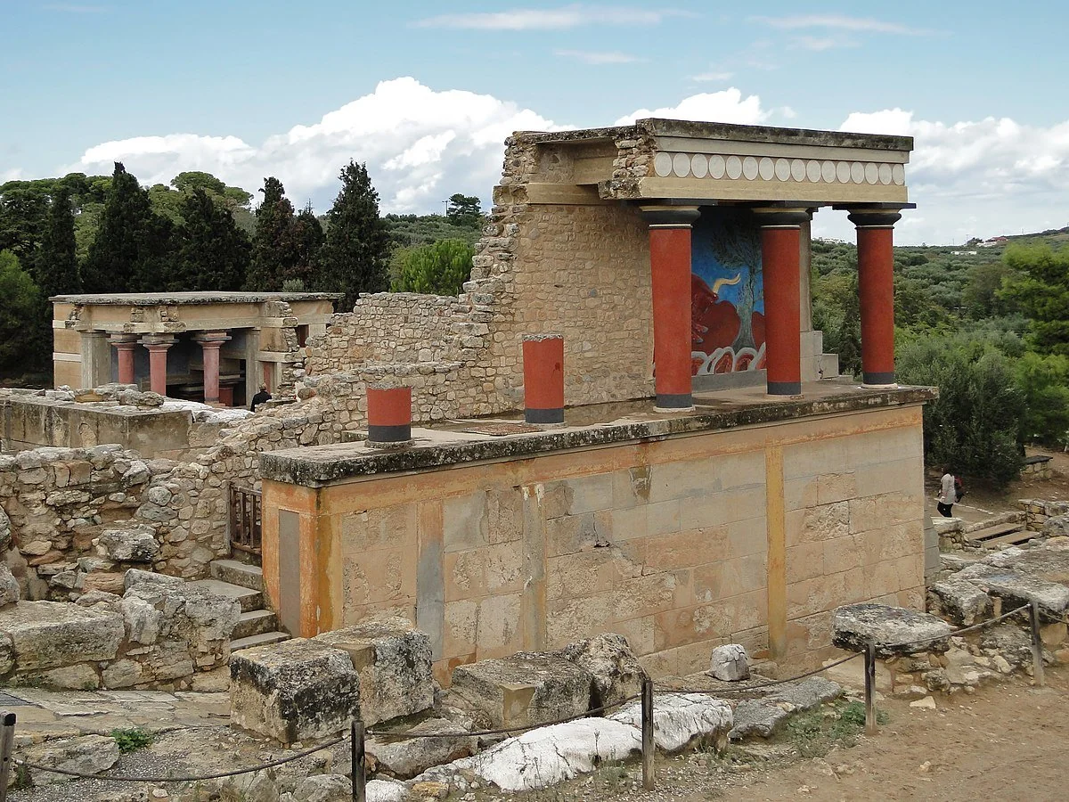 Ruins of the ancient Palace of Knossos in Crete, featuring partially restored stone walls, red columns, and a colorful fresco, surrounded by trees and scattered stones.