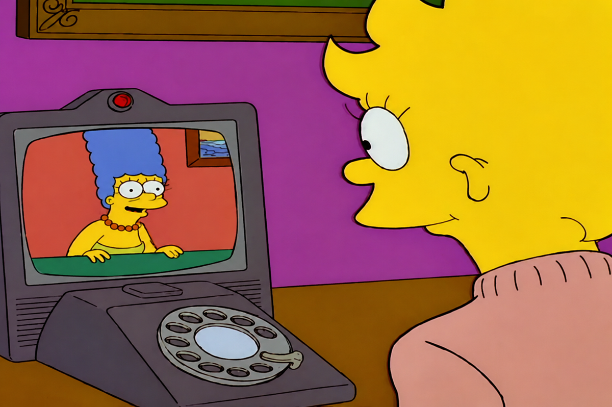 Lisa Simpson video chats with Marge Simpson, who appears on the screen of a retro videophone with a rotary dial. Lisa sits at a desk, and the walls are purple.