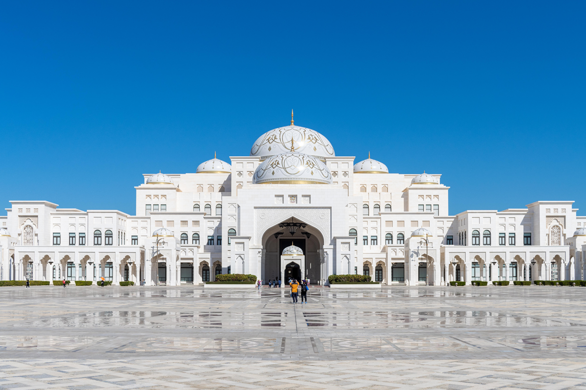 A grand white palace with ornate domes and arches stands under a clear blue sky; people walk towards its entrance across a spacious, patterned plaza.
