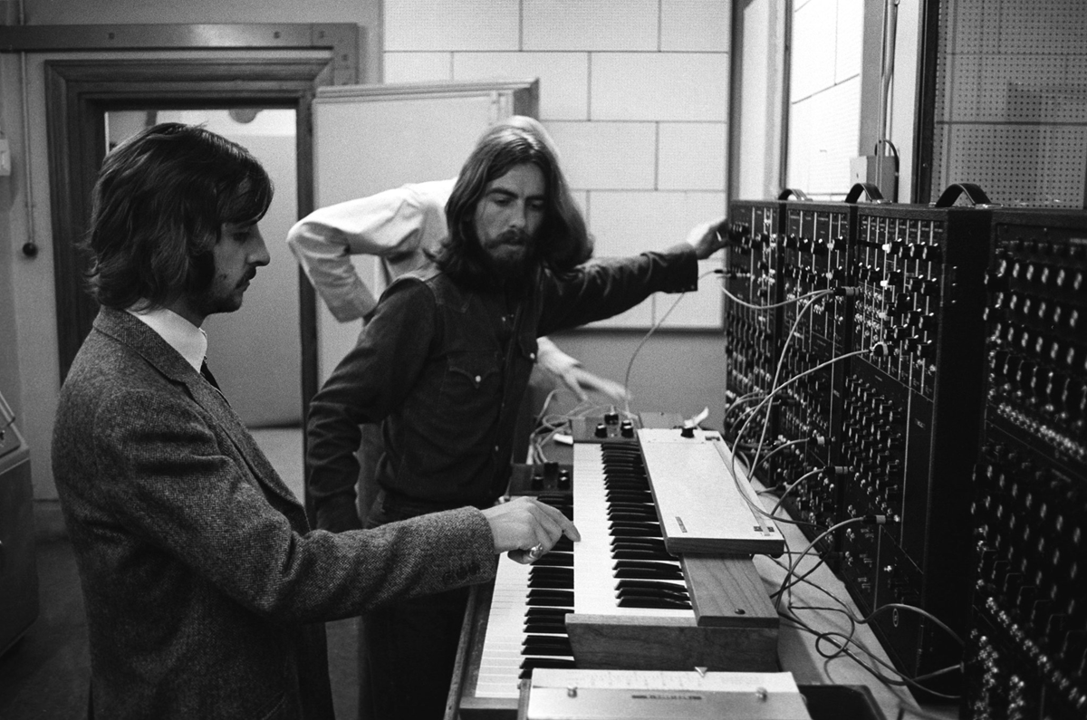Two men operate a large modular synthesizer with many cables and knobs; one plays a keyboard while the other adjusts the equipment. A third person stands behind them, partially visible. The scene is in a studio or lab setting.