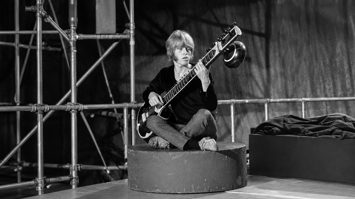 A young person with light hair sits cross-legged on a round platform, playing a sitar. Metal scaffolding and a draped cloth are visible in the background. The image is in black and white.